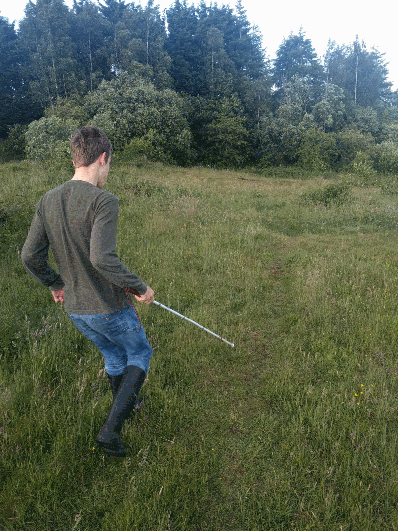 Ethan walks, with cane out front, over a grassy field.  His back is to the camera.
