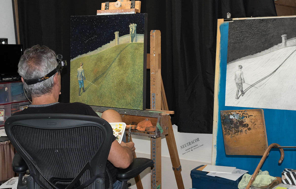 Artist Tony Luciani sits in front of a canvas with the front cover painting completed on it.  To his right is the pencil sketch on another easel.  Tony's back is to the camera and his sits contemplating the painting.